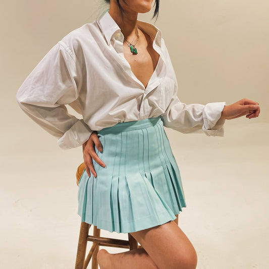 woman sitting on wood stool wearing oversized white button down and teal pleated tennis skirt and jade necklaces